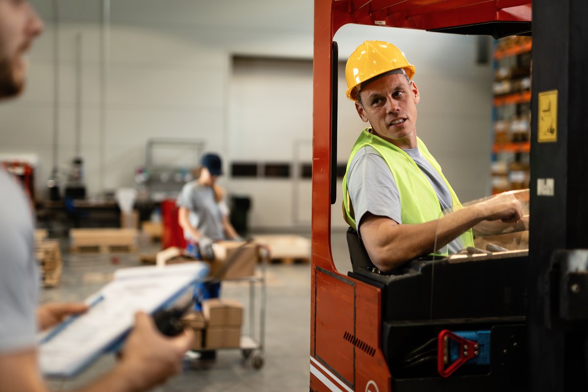 Smiling forklift driver communicating with dispatcher about cargo detail in industrial warehouse