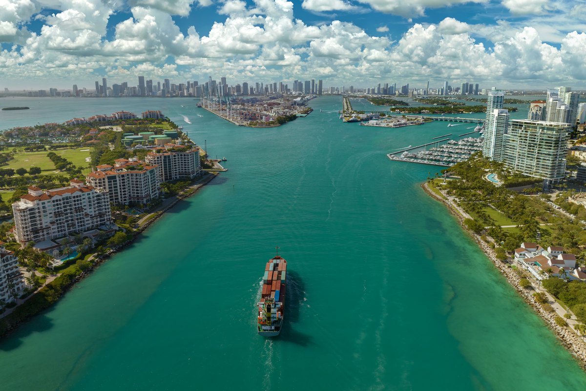 Aerial view of large container ship entering in miami harbor main channel near south beach luxurious hotels and apartment buildings on waterfront and high skyscrapers of downtown district in distance